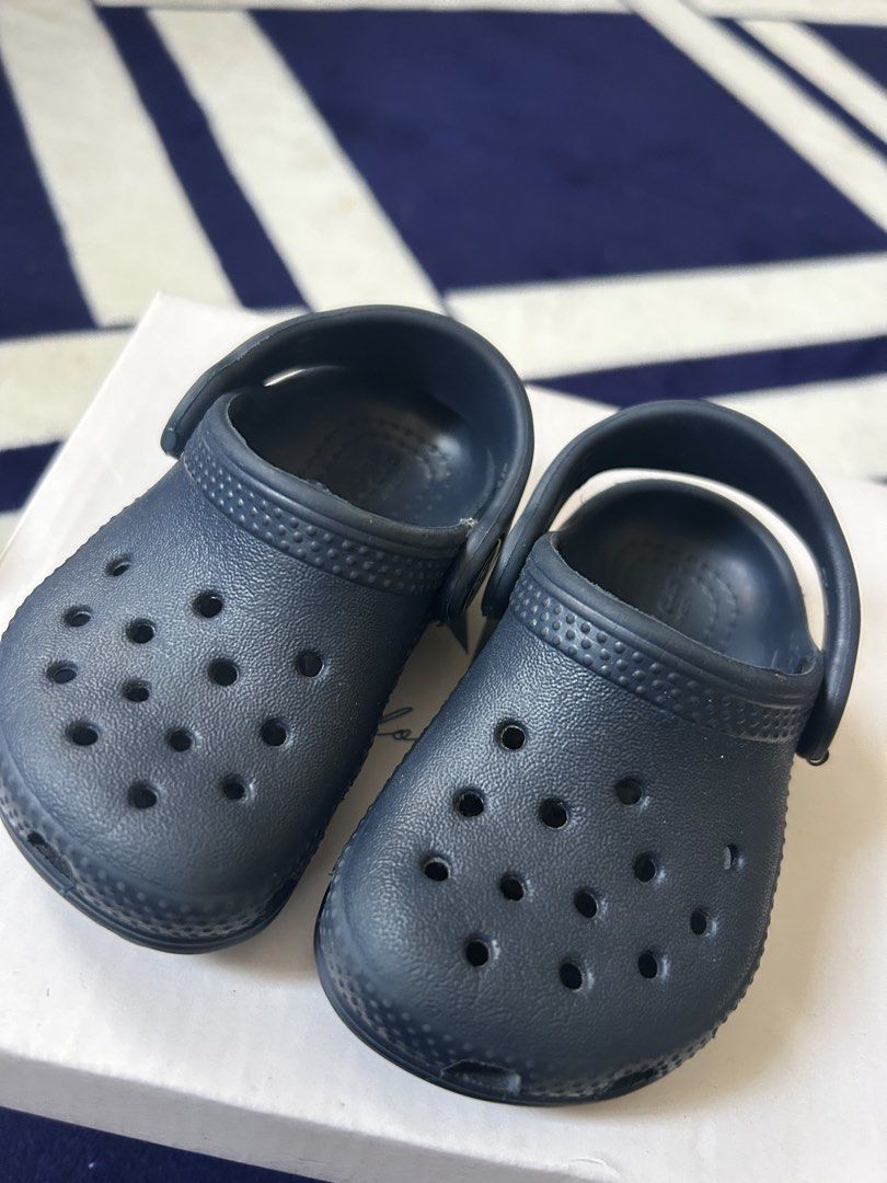 Crocs Navy Blue Toddler Clogs Size C4 (Category: Babies & Kids Fashion) located in Manila. Submitted by oreocat18 (Listing ID 1427683628, Image 1). Description: Authentic navy blue Crocs toddler clogs. Features the classic Crocs design with ventilation holes and a pivoting heel strap for a secure fit. The size is C4. The Crocs logo is visible on the side of each shoe. The soles show the Crocs branding and size information..