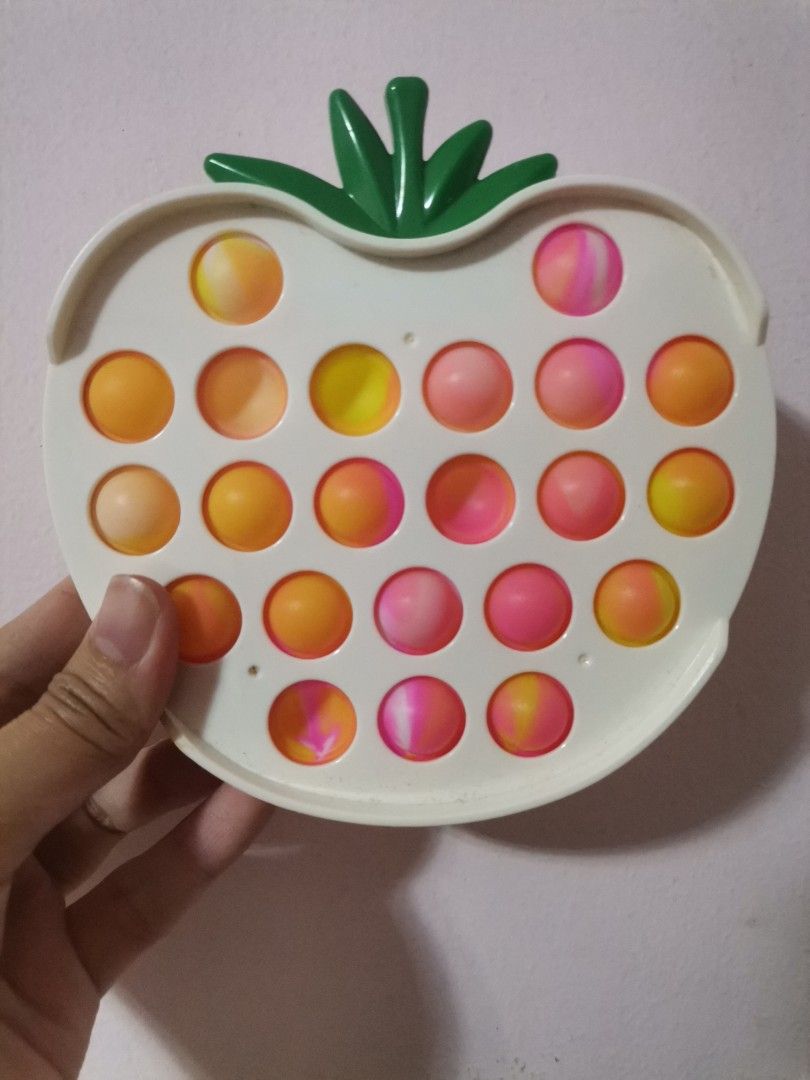 Strawberry Pop It Fidget Toy (Category: Kids Toys) located in Makati City. Submitted by kookybearsstuff (Listing ID 1427875996, Image 2). Description: Strawberry-shaped Pop It fidget toy. It has a white base with a green stem. The bubbles are a mix of orange, yellow, and pink. It is designed to provide sensory stimulation and stress relief. The toy is held in a hand in the images..