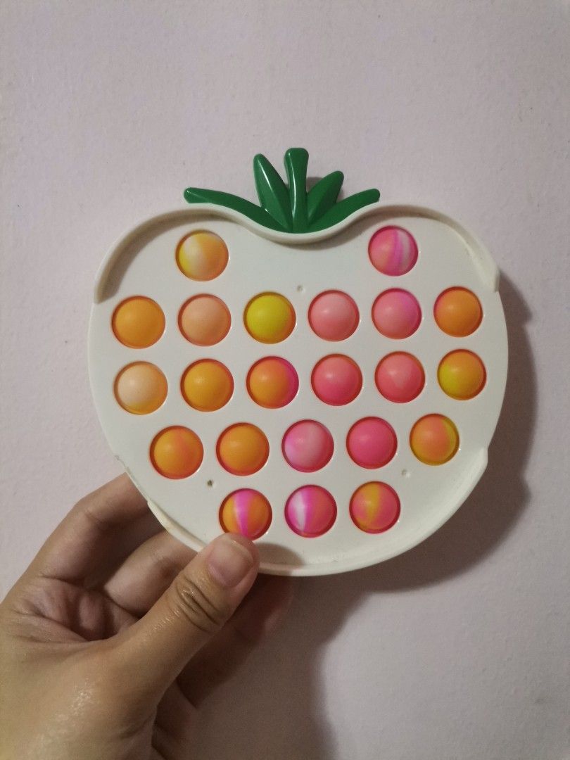 Strawberry Pop It Fidget Toy (Category: Kids Toys) located in Makati City. Submitted by kookybearsstuff (Listing ID 1427875996, Image 1). Description: Strawberry-shaped Pop It fidget toy. It has a white base with a green stem. The bubbles are a mix of orange, yellow, and pink. It is designed to provide sensory stimulation and stress relief. The toy is held in a hand in the images..