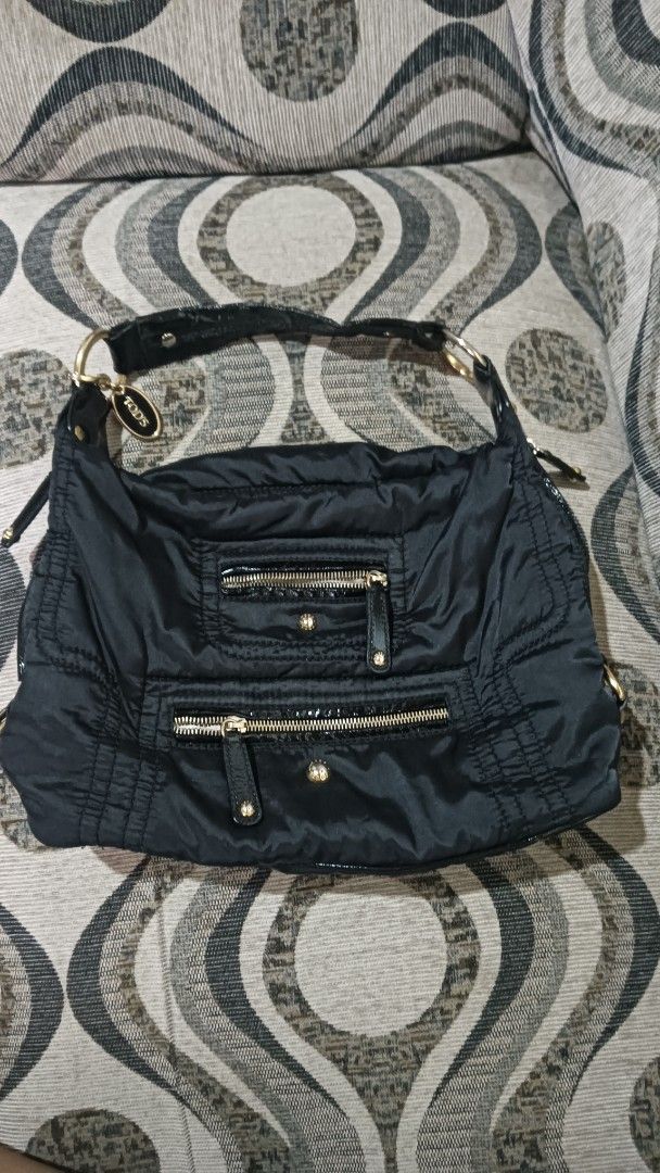 Tod's Black Nylon Handbag (Category: Luxury Bags & Wallets) located in Laoag. Submitted by catrnzjb153008 (Listing ID 1427823626, Image 1). Description: Black nylon handbag from Tod's. Features multiple zippered compartments and gold-toned hardware. The interior lining is a light, silky material. The bag has a shoulder strap with a patent leather accent. The Tod's logo is visible on the zipper and the strap. Includes a small charm with the Tod's 