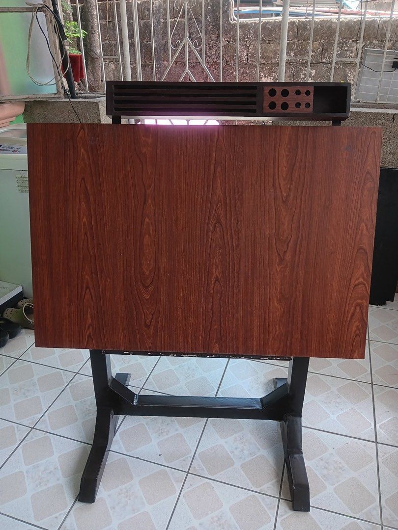 Wooden Drafting Table with Storage (Category: Tables & Sets) located in Pasig City. Submitted by khiarha (Listing ID 1427985217, Image 2). Description: Wooden drafting table features a dark wood finish and a built-in storage compartment. It has a slatted top section and a section with multiple circular holes. The table is supported by a sturdy metal frame. The table's surface has a wood grain pattern. The table also has a storage area underneath 