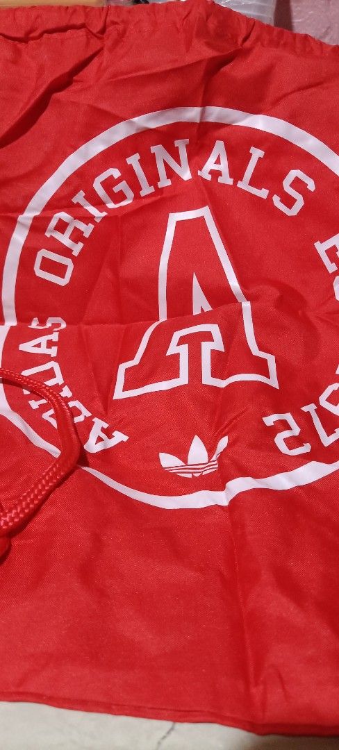 Adidas Originals Drawstring Bag (Category: Backpacks) located in Bocaue. Submitted by michellesell_2 (Listing ID 1428228822, Image 1). Description: Red drawstring bag featuring the Adidas Originals logo. The bag has a large white circular design with the word "ORIGINALS" and a large "A" in the center. The Adidas trefoil logo is visible below the "A"..