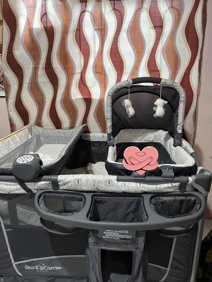 Graco Pack 'n Play Playard with Bassinet (Category: Cots & Cribs) located in San Fernando. Submitted by c.amerrie (Listing ID 1428928433, Image 1). Description: Graco Pack 'n Play playard includes a bassinet and a changing station. It features a mobile with hanging toys and storage pockets. The playard has a gray and white design with patterned fabric..