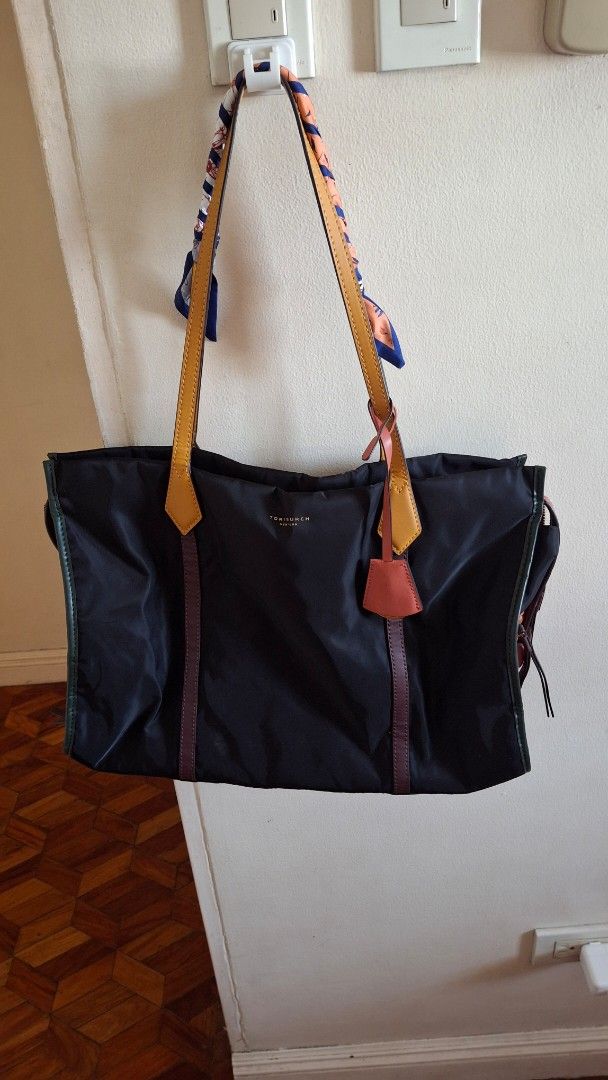 ORIGINAL Tory Burch Nylon Tote Bag (Category: Luxury Bags & Wallets) located in Las Pinas. Submitted by mgr29 (Listing ID 1429172813, Image 1). Description: FLAWS: DOG BITE - USED ELECTRIC TAPE TO HIDE AND USED A SCARF TO WRAP THE HANDLE. SLIGHT PEELING IN ONE OF THE HANDLES USED TWICE AND STORED FOR A LONG TIEM. Black nylon tote bag from Tory Burch. Features leather straps and accents in brown and orange. Includes a patterned scarf tied to the h