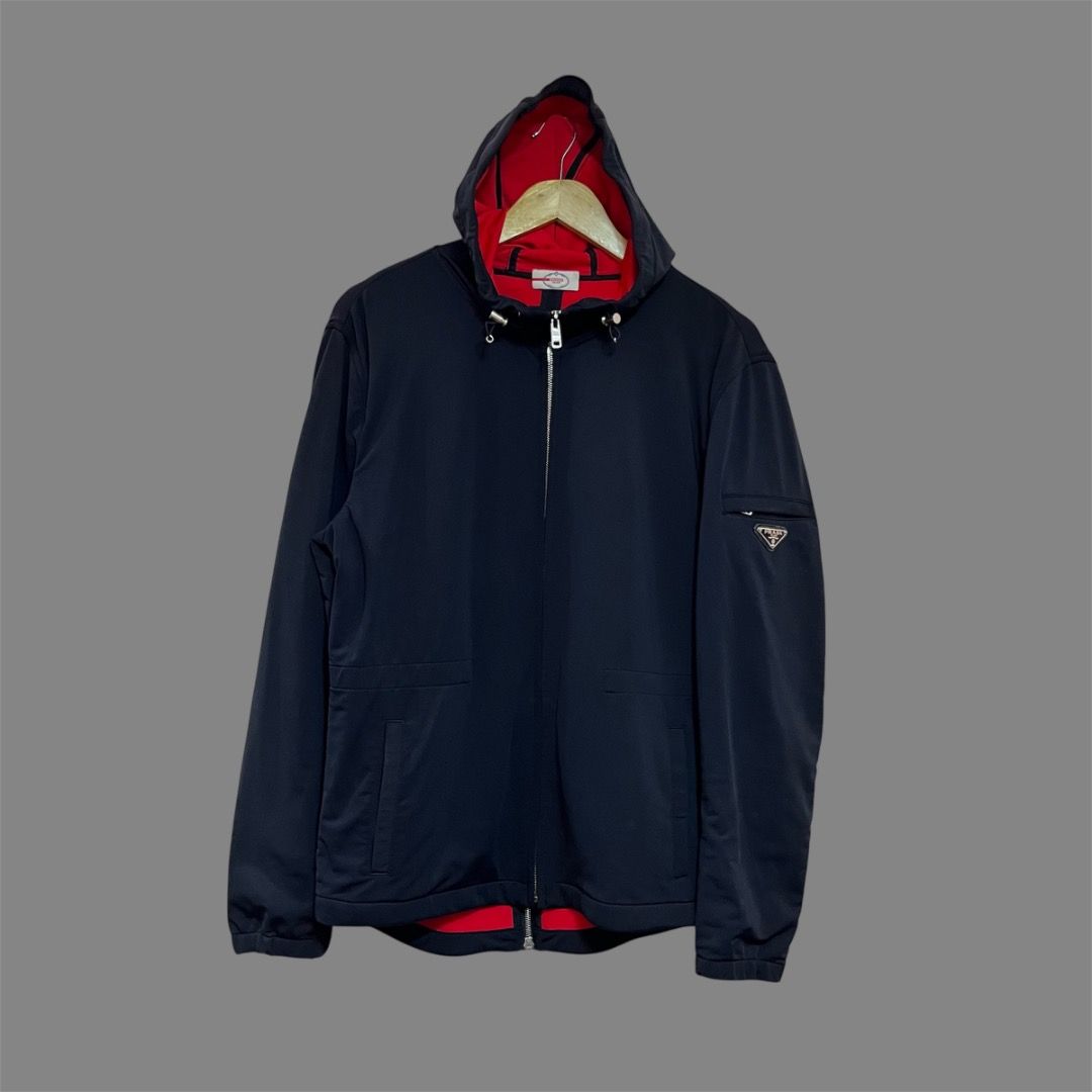 Prada Sport Jacket - Black and Red Lining (Category: Luxury Apparel) located in Bacolod City. Submitted by boujee.wardrobes (Listing ID 1429319991, Image 1). Description: Prada hooded jacket. Features a black exterior with a red interior lining. Includes a zip closure, a Prada logo on the sleeve, and a size tag inside. The jacket has a hood with adjustable drawstrings and a zip pocket. Size is Tg.50. 27L 21.5W.