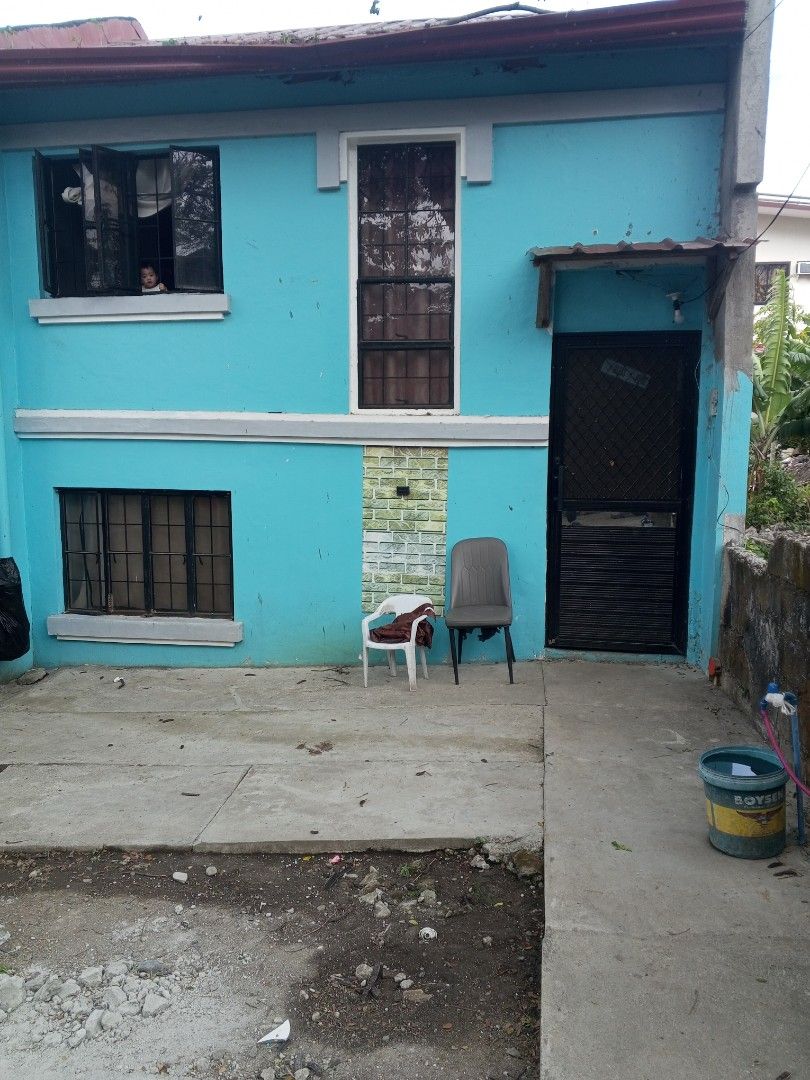 Two-Story House with Blue Exterior (Category: House & Lot) located in Trece Martires City. Submitted by djaothriftshop (Listing ID 1429266722, Image 1). Description: Two-story house with a blue exterior. Features include multiple windows, a black door, and a small porch area. There is a small brick accent near the center window. Two chairs are placed on the porch. The house has a concrete pathway leading to the entrance..