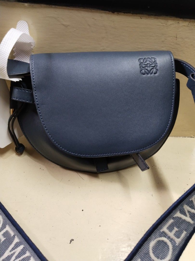 Loewe Gate Dual Mini Bag (Category: Luxury Bags & Wallets) located in Baguio. Submitted by bangtowanv91137 (Listing ID 1424604042, Image 1). Description: Loewe Gate Dual Mini Bag features a navy blue leather exterior with a matching strap. The bag has a saddle shape with the Loewe logo embossed on the front. The interior is lined with a tan suede-like material and includes a small pocket. The strap is a woven design with the Loewe logo..