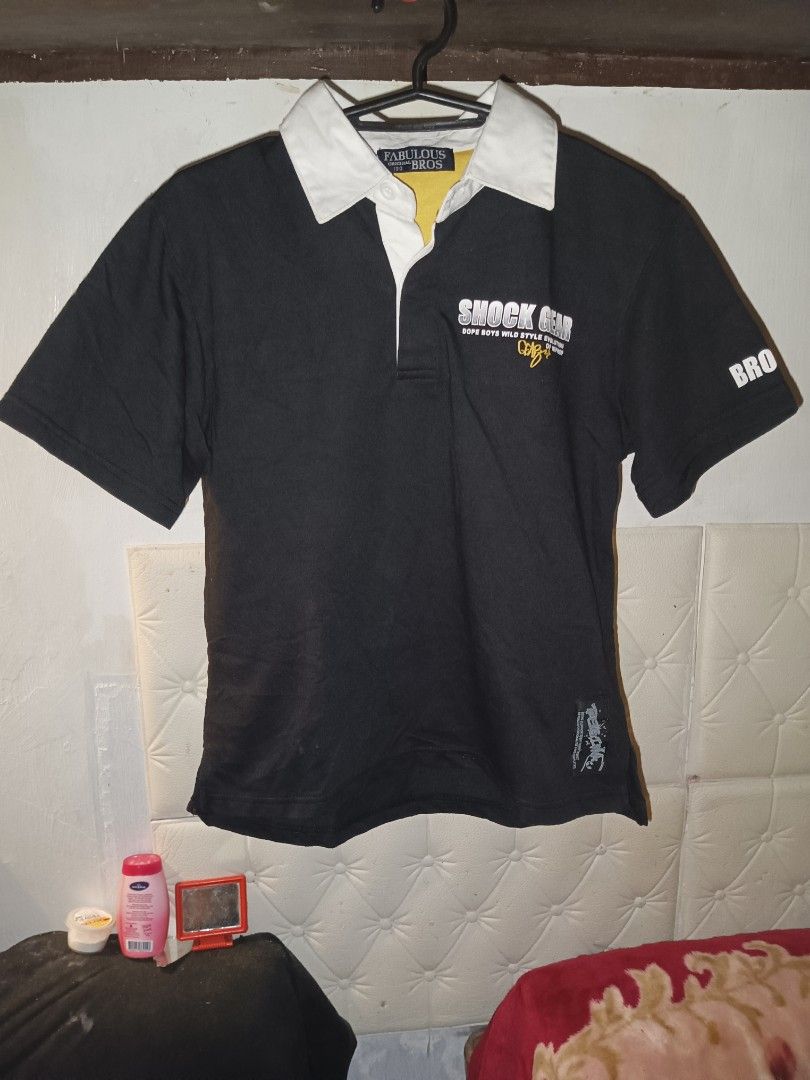 Shock Gear Rugby Shirt (Category: Tshirts & Polo Shirts ) located in Iloilo. Submitted by junpopakoto (Listing ID 1424542770, Image 1). Description: Black rugby shirt with white collar and yellow inner collar. Features "Shock Gear" graphic on the front and a skater graphic with text on the back. Short-sleeved with a button-up collar. The shirt has a "BRO" print on the right sleeve and "OKLIN" on the left sleeve..