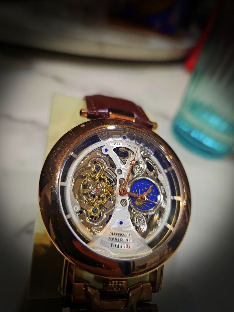 Difful Original Automatic Skeleton Watch (Category: Watches) located in Kuala Lumpur. Submitted by freewhale-6bcf (Listing ID 1424866061, Image 1). Description: Difful Original Automatic watch features a skeleton design, showcasing the intricate inner workings. It has a brown leather strap and a gold-colored case. The watch also has a moon phase indicator. The back of the watch is transparent, revealing the automatic movement. The clasp is brande