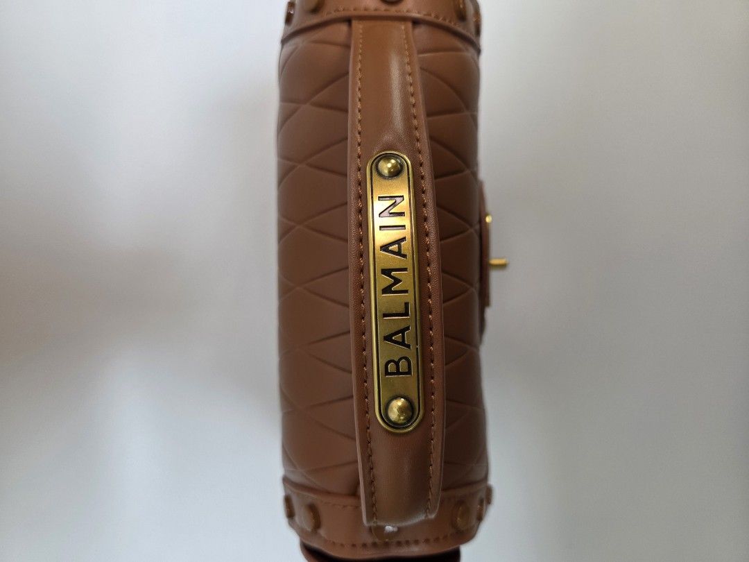 Balmain Brown Crossbody Bag (Category: Luxury Bags & Wallets) located in Quezon City. Submitted by robyns.closet.ph (Listing ID 1425411038, Image 3). Description: Brown crossbody bag by Balmain. Features a structured design with a top handle and a detachable shoulder strap. The bag has a flap closure and a gold-tone logo on the front. Interior includes a zippered pocket. Detailed stitching and rivet accents add to the bag's aesthetic..