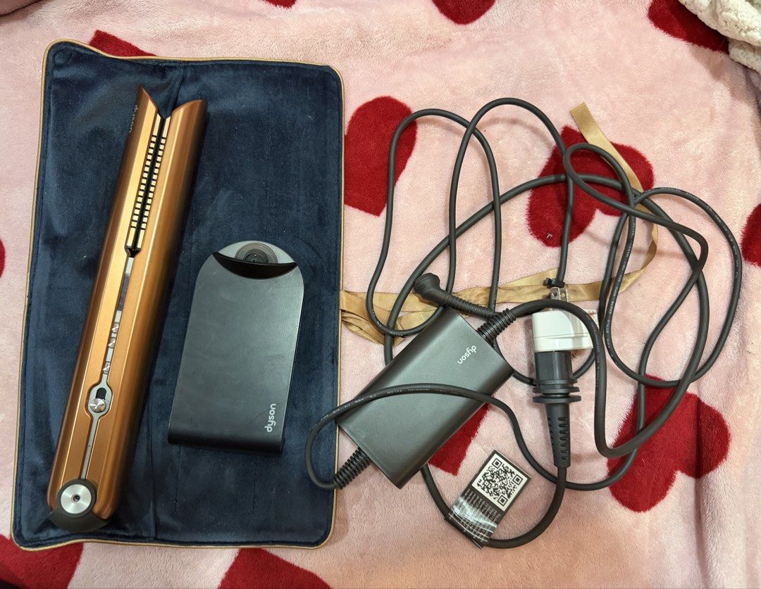 Dyson Corrale Hair Straightener (Category: Hair) located in Tangub. Submitted by pollymolly23 (Listing ID 1425376688, Image 1). Description: Dyson Corrale hair straightener in copper color. Includes charging dock and charger. The straightener is placed on a blue velvet-like mat. The charger has a QR code attached. The power cord is black..