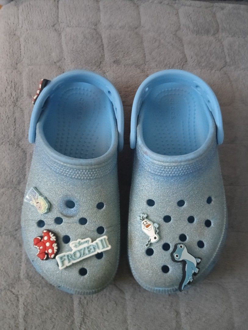 Crocs Frozen Jibbitz Clogs (Category: Babies & Kids Fashion) located in Santa Rosa. Submitted by amiraamara (Listing ID 1425499087, Image 1). Description: Light blue Crocs clogs with Frozen-themed Jibbitz. Features a glittery finish and a raised sole. Includes Jibbitz of Olaf, Elsa, and other Frozen characters. The size is J1. The Crocs logo is visible on the side and sole of the shoes. The heel strap has the Crocs logo..