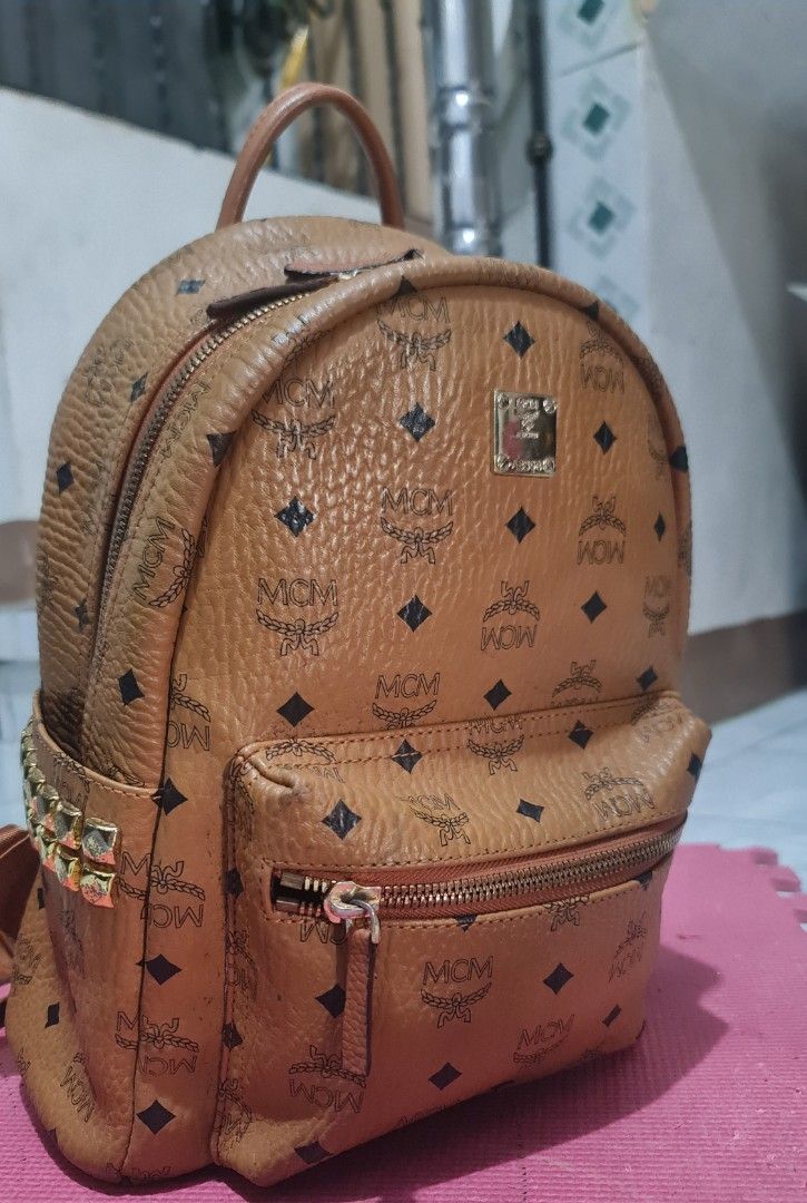 Authentic MCM Backpack (Category: Luxury Bags & Wallets) located in Lucena. Submitted by gwenbibi17 (Listing ID 1428993436, Image 1). Description: Brown MCM backpack with a front zippered pocket. Features the MCM logo pattern and a small metal logo plate. The backpack has gold-colored hardware and studded accents..
