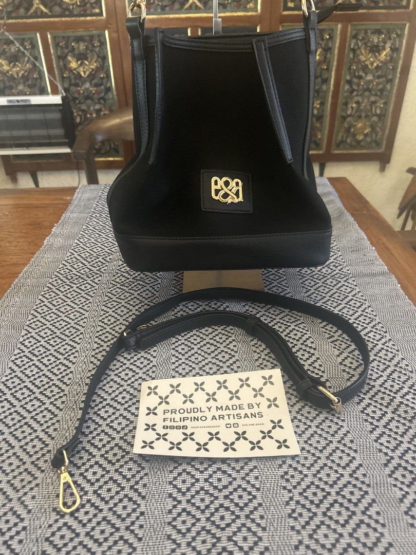 Eye and Adam Black Handbag (Category: Luxury Bags & Wallets) located in Las Pinas. Submitted by tinamontec00246 (Listing ID 1431821783, Image 3). Description: Black handbag with a gold logo. Includes a detachable shoulder strap. The bag has a structured shape with a flat base. The interior has multiple compartments. The bag is proudly made by Filipino artisans. L-10” W- 10&1/2” Diameter -6” Handle drop -8” Comes with long adjustable shoulder stra