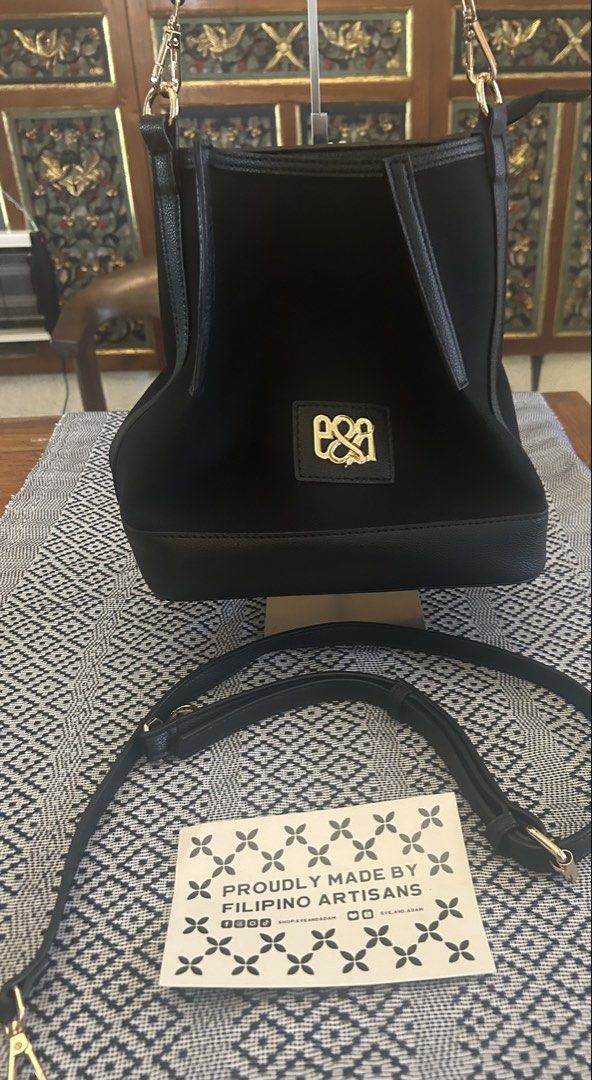 Eye and Adam Black Handbag (Category: Luxury Bags & Wallets) located in Las Pinas. Submitted by tinamontec00246 (Listing ID 1431821783, Image 1). Description: Black handbag with a gold logo. Includes a detachable shoulder strap. The bag has a structured shape with a flat base. The interior has multiple compartments. The bag is proudly made by Filipino artisans. L-10” W- 10&1/2” Diameter -6” Handle drop -8” Comes with long adjustable shoulder stra
