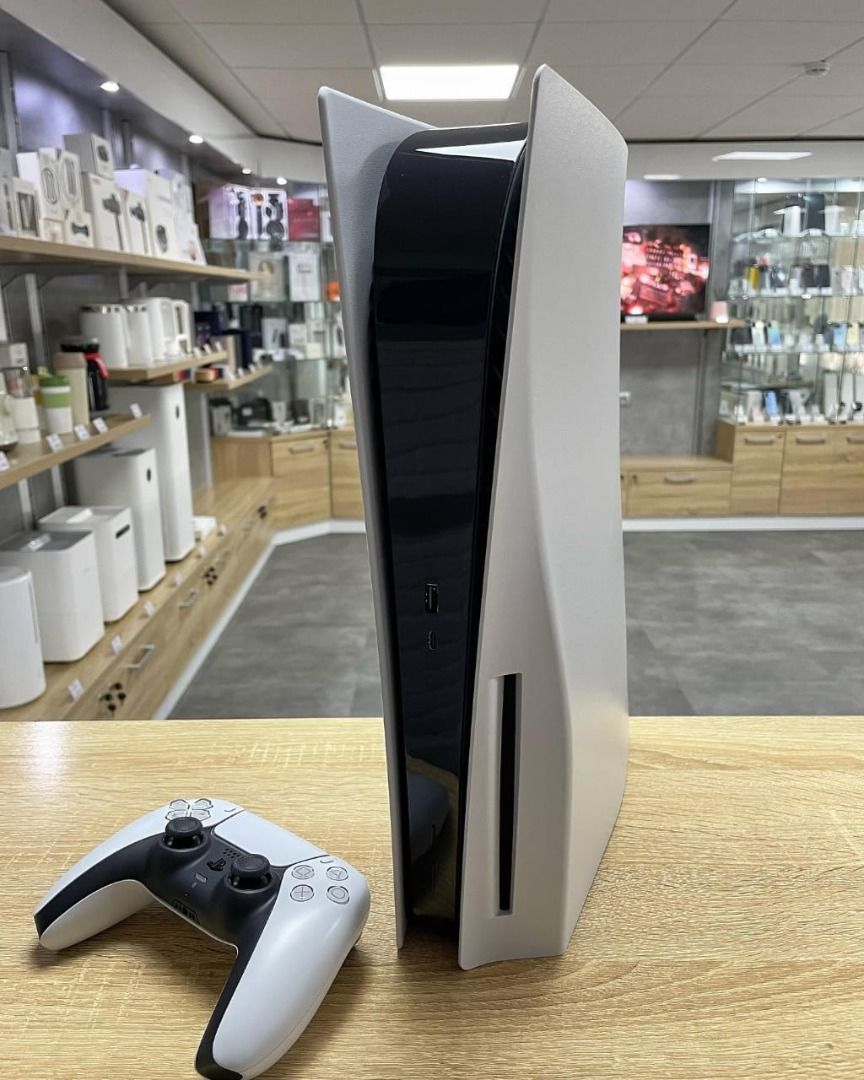 Sony PlayStation 5 Console (Category: PlayStation) located in Caloocan City. Submitted by truehawk.889b68 (Listing ID 1432027994, Image 2). Description: PlayStation 5 console is shown with a white and black design. It includes a white DualSense controller. The console is standing upright on a wooden surface. The controller is also placed on the wooden surface. The console has a sleek, modern aesthetic..