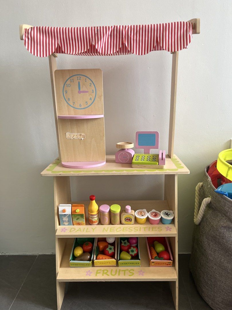 Wooden Play Shop Stall (Category: Infant Playtime) located in Cainta. Submitted by danica081434855 (Listing ID 1432223355, Image 1). Description: - Wooden play shop stall - Features a clock, shelves (Daily Necessities & Fruits), and striped awning - Includes toy food items - Comes with a cash register - Encourages imaginative play - Perfect for kids who love to pretend shop.
