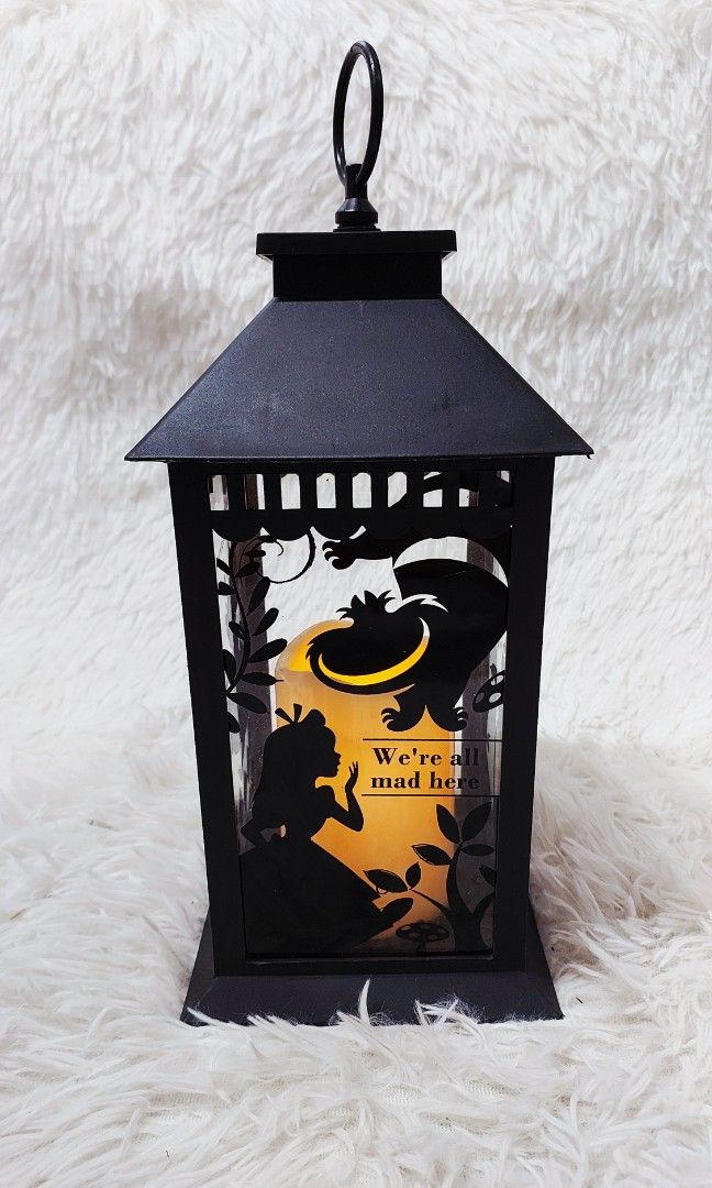 Alice in Wonderland-Inspired Lantern (Category: Lighting) located in Binan City. Submitted by sakuratreasuresph (Listing ID 1432698822, Image 2). Description: Fall down the rabbit hole into a magical glow with this Alice in Wonderland-inspired lantern 🐇✨🖤 Featuring a charming silhouette of Alice and the Cheshire Cat 😸, this lantern lights up with a warm, cozy glow that brings a dreamy, storybook vibe to any space. The iconic phrase “We’re all 