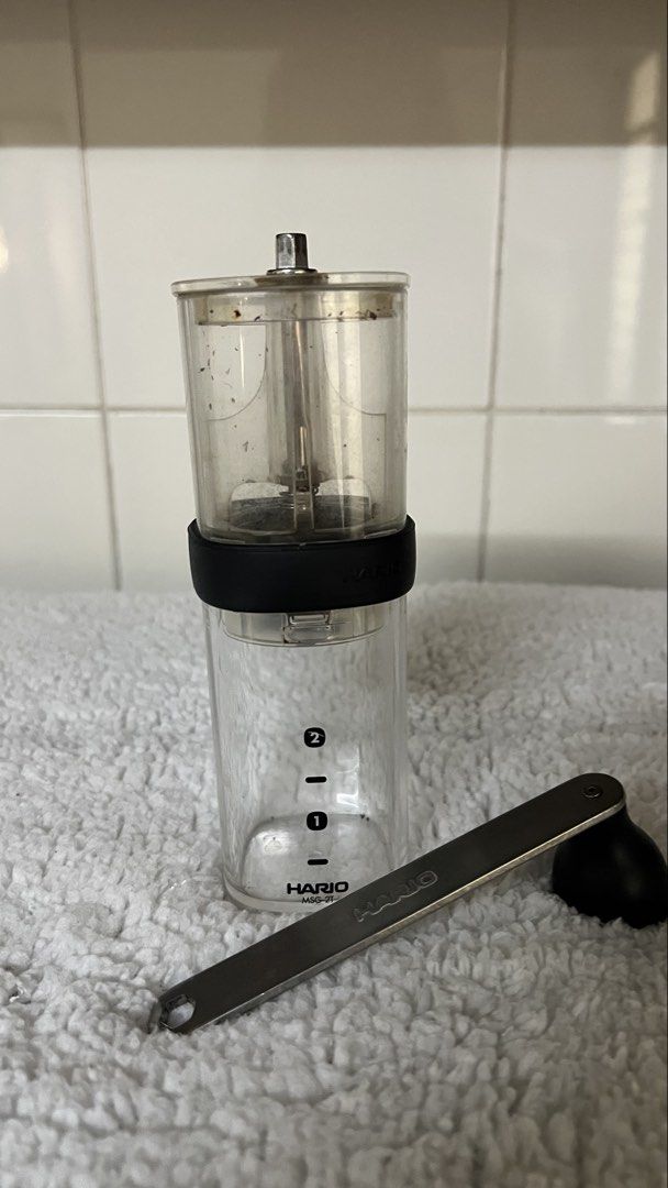 Hario Coffee Grinder (Category: Coffee Machines & Makers) located in Santa Ana. Submitted by hamburjei_thrifts (Listing ID 1433289225, Image 1). Description: - Hario coffee grinder - Includes grinder and measuring spoon - Clear glass container with measurement markings - Model number visible on spoon.