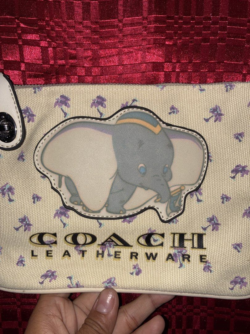 Coach Dumbo Leatherware Clutch (Category: Luxury Bags & Wallets) located in Placer. Submitted by talaalond61563 (Listing ID 1433782248, Image 2). Description: - Authentic Coach Dumbo clutch - Features a charming Dumbo design - Cream-colored background with purple floral accents - Secure zipper closure - Includes a convenient wrist strap - Interior: Coach label with serial number H1777-25617 - Exterior: Coach Leatherware logo.