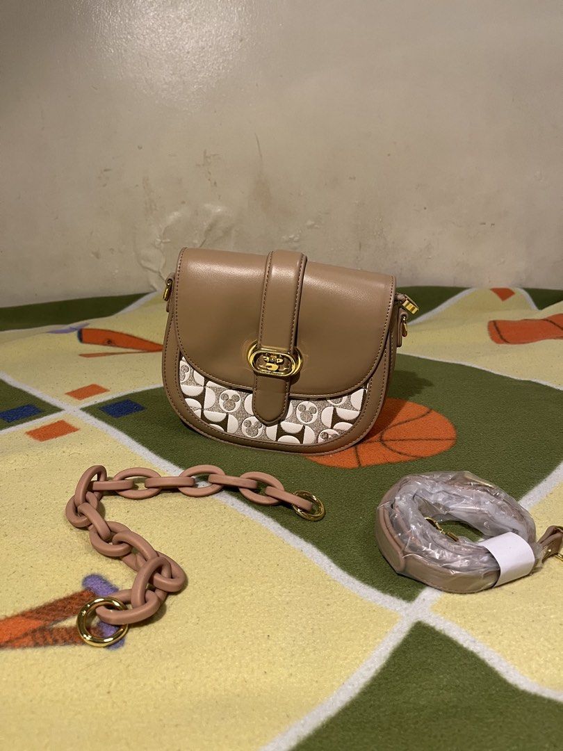 Crossbag saddle bag (DISNEY X COACH) (Category: Luxury Bags & Wallets) located in Caloocan City. Submitted by lawrence.jay (Listing ID 1433885300, Image 3). Description: Coach saddle bag features a unique Mickey Mouse pattern on the front. It has a tan leather exterior with a gold-tone clasp and hardware. The bag comes with a detachable shoulder strap and a chain strap. The design is a collaboration with Disney..