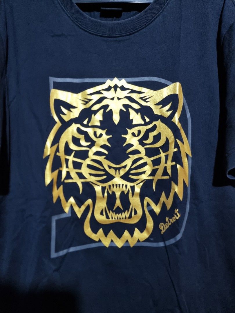 MLB Detroit Tigers Graphic Tee - Large (Category: Tshirts & Polo Shirts ) located in Imus. Submitted by aim_for_less (Listing ID 1434117562, Image 3). Description: MLB Detroit Tigers Graphic Tee Features a striking gold tiger design on a navy blue background. It has a crew neck and short sleeves. The tag indicates it is a size Large (175/92A). Made by MLB (Major League Baseball) -DIMES: 21" X 29" - COLOR RATE: 9.7/10 - NO ISSUE - VERY GOOD CONDIT