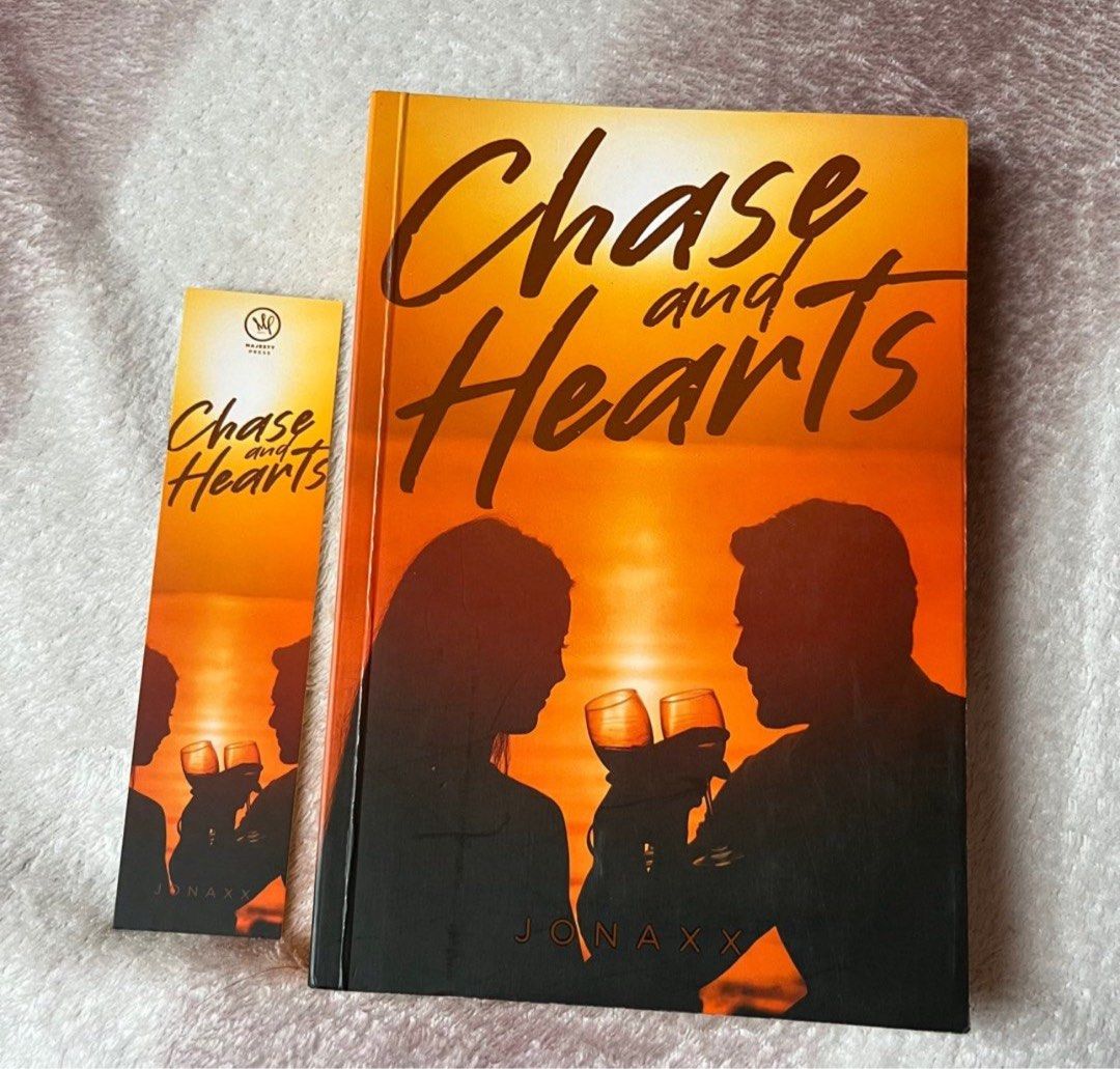 Chase and Hearts by Jonaxx (Category: Storybooks) located in Manila. Submitted by chicdistrict.ph (Listing ID 1434356103, Image 1). Description: - "Chase and Hearts" by Jonaxx book included. - Comes with a matching bookmark. - Cover art: Silhouette of a couple toasting with wine glasses against an orange sunset. - Author's name, Jonaxx, is on the cover..