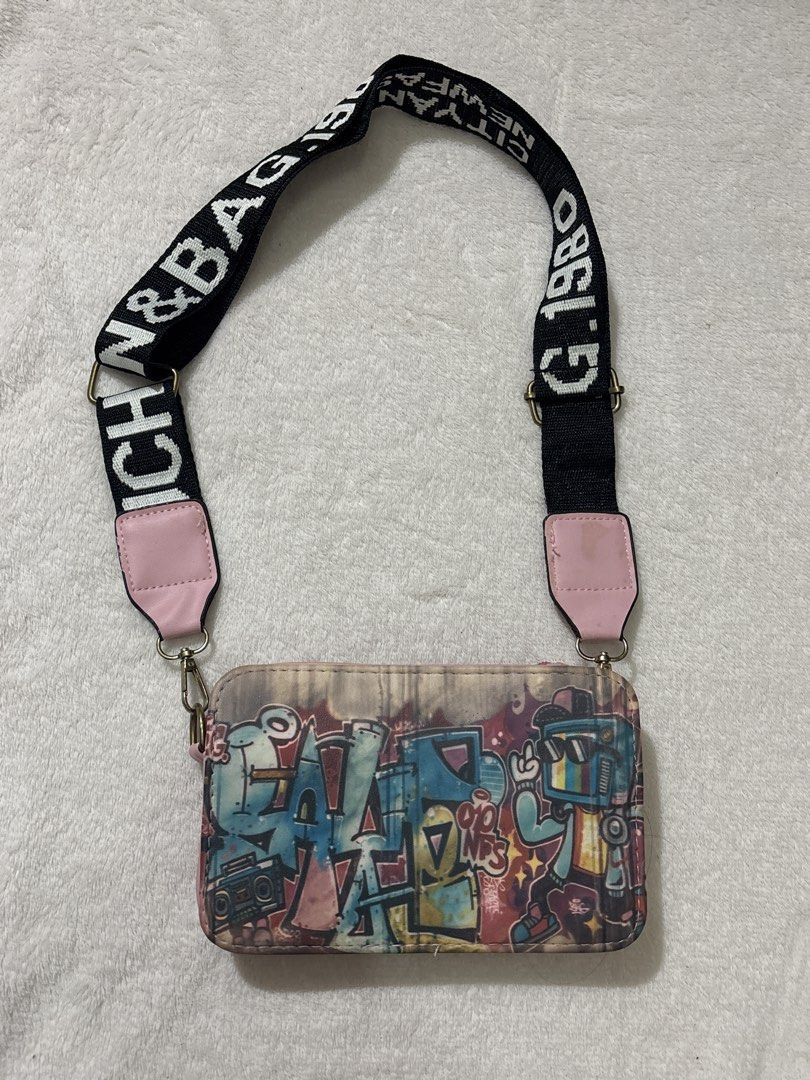 Graffiti Print Crossbody Bag (Category: Cross-body Bags) located in Muntinlupa City. Submitted by findsbymina (Listing ID 1434362439, Image 1). Description: Original Price: Approximately P700 Measurements: Length: 7.5 inches Width: 4.7 inches Description: This is a Bench Graffiti Street Art Crossbody Bag. It features a “guitar strap” style handle, which was a major trend in the mid-to-late 2010s and has recently made a comeback with the Y2K/Retro
