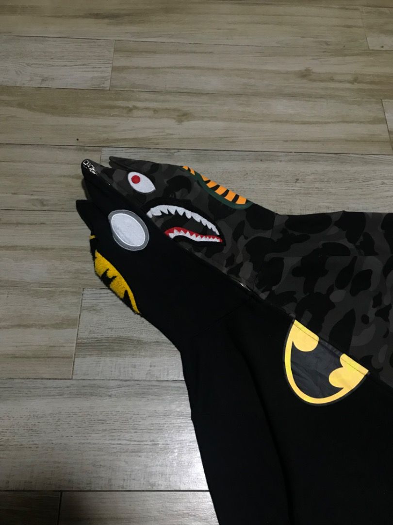 BAPE Batman Shark Hoodie (Category: Coats, Jackets and Outerwear) located in Manila. Submitted by timiasunci72128 (Listing ID 1434785351, Image 3). Description: BAPE hoodie features a unique split design with a Batman logo on one side and a shark face on the other. It has a full zip closure and a hood with ears. The hoodie showcases a camouflage pattern on the shark side and a yellow Batman logo. The DC logo is visible on the sleeve..