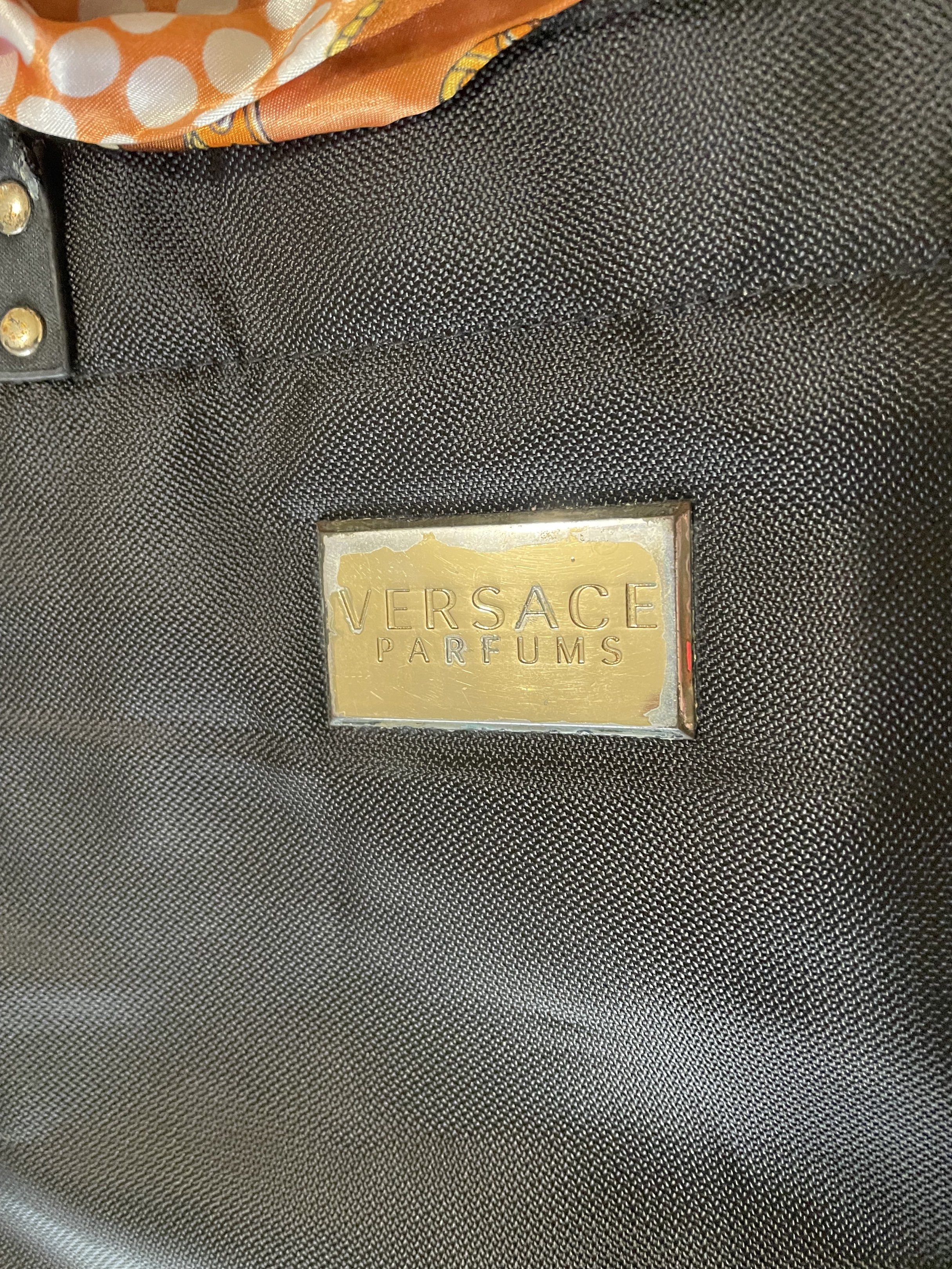 Versace Parfums Black Tote Bag (Category: Luxury Bags & Wallets) located in Santa Barbara. Submitted by frryt (Listing ID 1434603027, Image 2). Description: - Versace Parfums black tote bag - Gold Versace logo on front - Gold and black patterned accents - Includes matching scarf - Interior features gold and black Versace pattern - Zippered pocket with Versace Parfums zipper pull.