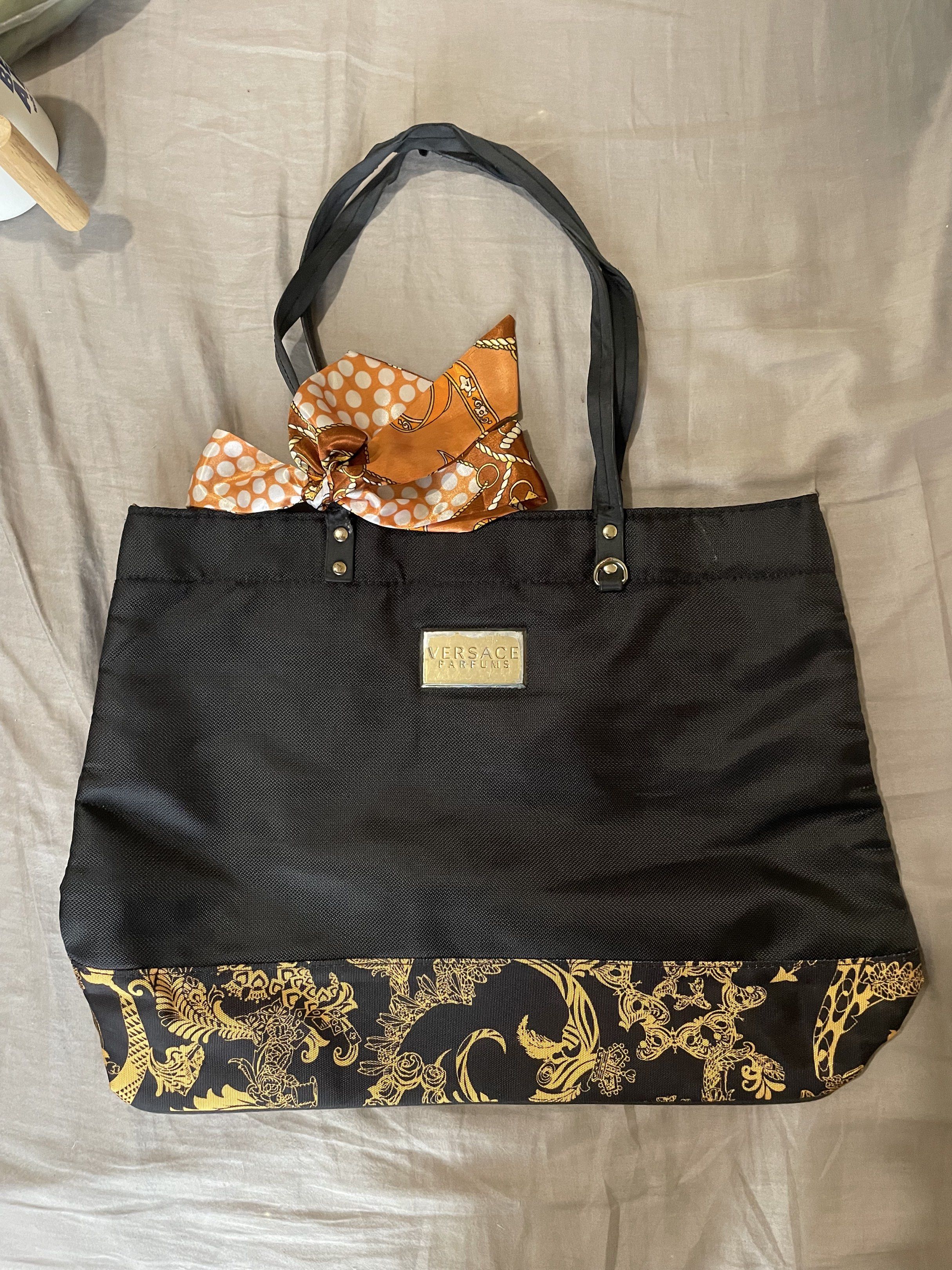 Versace Parfums Black Tote Bag (Category: Luxury Bags & Wallets) located in Santa Barbara. Submitted by frryt (Listing ID 1434603027, Image 1). Description: - Versace Parfums black tote bag - Gold Versace logo on front - Gold and black patterned accents - Includes matching scarf - Interior features gold and black Versace pattern - Zippered pocket with Versace Parfums zipper pull.