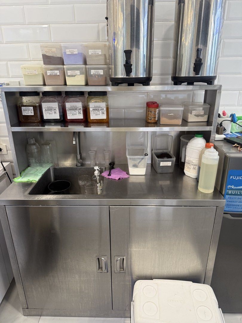 Stainless Steel Commercial Kitchen Sink (Category: Bathroom & Kitchen Fixtures) located in Manila. Submitted by alendlr (Listing ID 1434914449, Image 1). Description: - Commercial stainless steel kitchen sink with cabinet and shelving. - Includes two large beverage dispensers. - Comes with various ingredient containers. - Features a small sink area. - Cabinet with two doors for storage. - Ideal for small businesses or food service. .