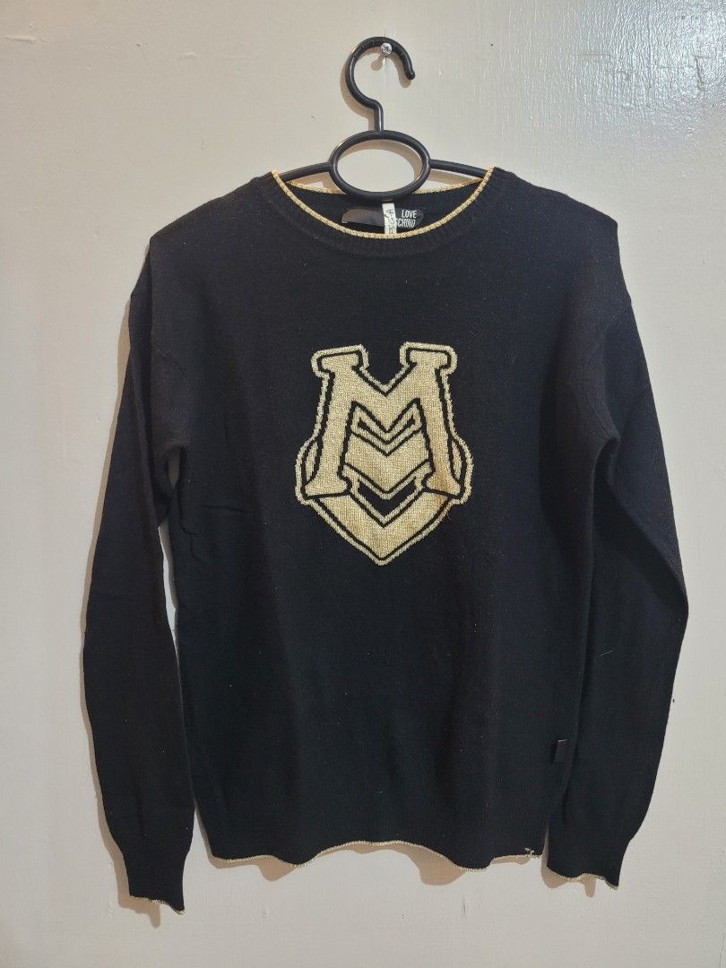 Love Moschino Black Sweater (Category: Coats, Jackets and Outerwear) located in Valenzuela. Submitted by flzoneeee (Listing ID 1429959421, Image 1). Description: Black sweater from Love Moschino. Features a large logo on the front. Made in Romania. The sweater has a round neckline with gold trim. The sleeves are long. The tag is visible, showing the brand name. The sweater is black..