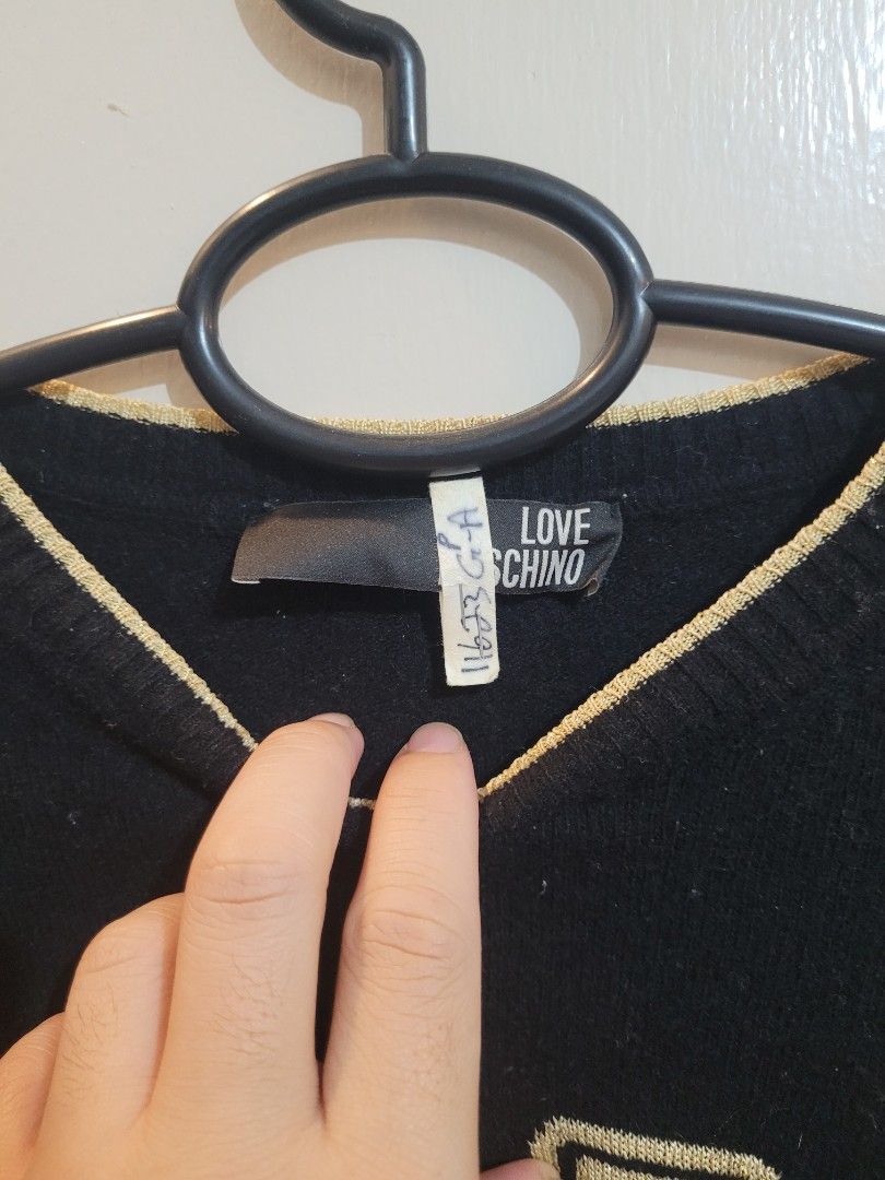Love Moschino Black Sweater (Category: Coats, Jackets and Outerwear) located in Valenzuela. Submitted by flzoneeee (Listing ID 1429959421, Image 3). Description: Black sweater from Love Moschino. Features a large logo on the front. Made in Romania. The sweater has a round neckline with gold trim. The sleeves are long. The tag is visible, showing the brand name. The sweater is black..