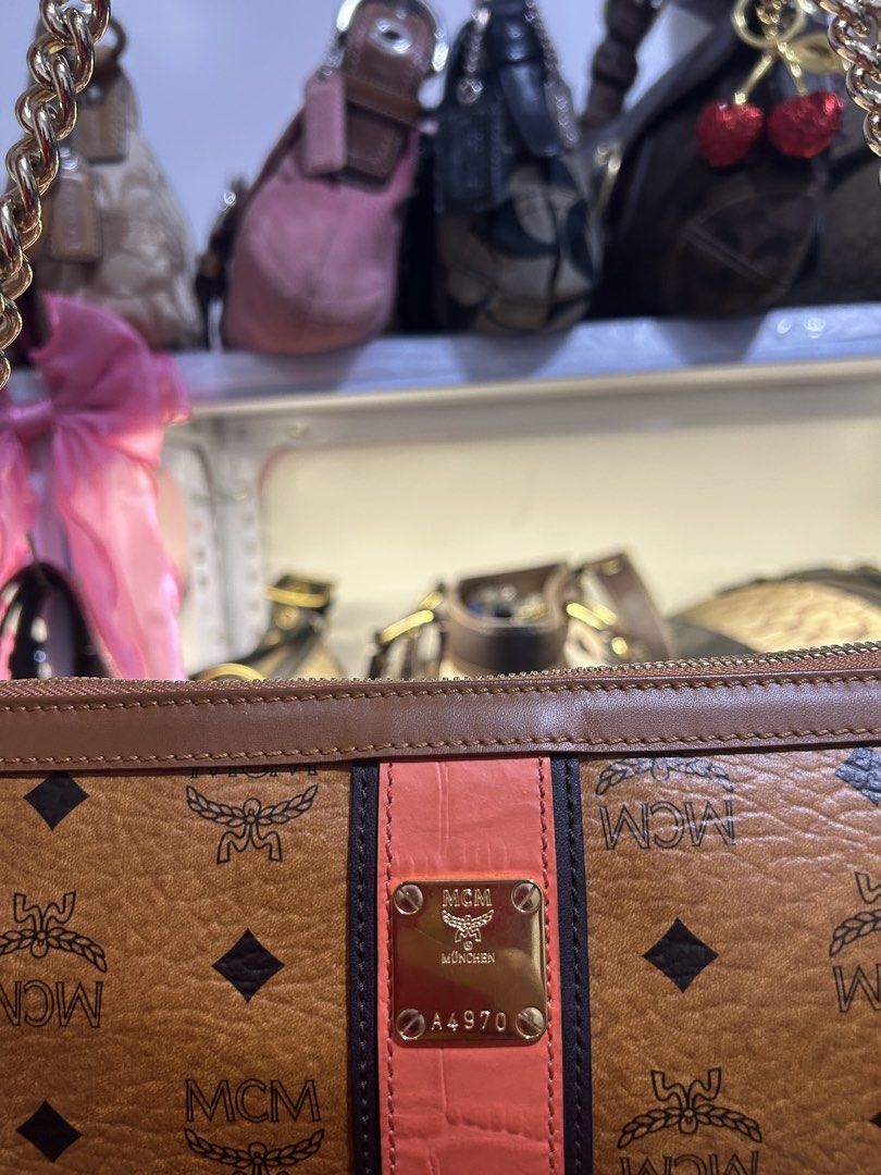 MCM Shoulder Bag with Chain (Category: Luxury Bags & Wallets) located in Manila. Submitted by graceyabut0 (Listing ID 1429949542, Image 2). Description: MCM shoulder bag features a brown monogram pattern with a pink stripe detail. It has a gold chain strap and a cherry charm. The interior includes multiple compartments and a zippered pocket. The bag is marked "Made in Korea" and has the MCM logo inside. The bag has a gold zipper and a gold MCM lo