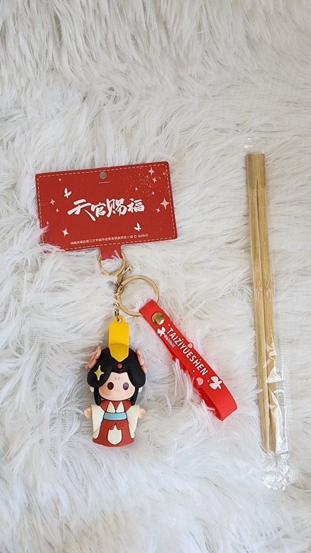 TGCF Crown Prince XieLian Keychain (Category: Character Goods) located in Manila. Submitted by quickocean (Listing ID 1430272757, Image 2). Description: Keychain features a cute character from Heaven Official's Blessing. It comes with a red strap that says "TAIZIYUESHEN". Also included are a red tag and a pair of chopsticks. The keychain is attached to a gold-colored ring. The character is wearing a red and white outfit with a yellow headpiece..