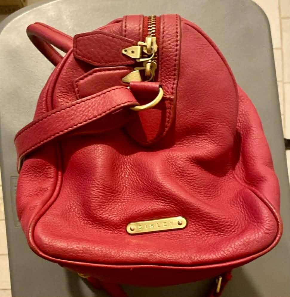 Sisley Red Leather Handbag (Category: Luxury Bags & Wallets) located in Marikina City. Submitted by imwondermum (Listing ID 1430704659, Image 2). Description: Red leather handbag from Sisley features a top handle and a detachable shoulder strap. It has a zip closure and gold-tone hardware. The interior is lined with red fabric and includes a small zippered pocket. The bag has a tag with the Sisley logo inside..