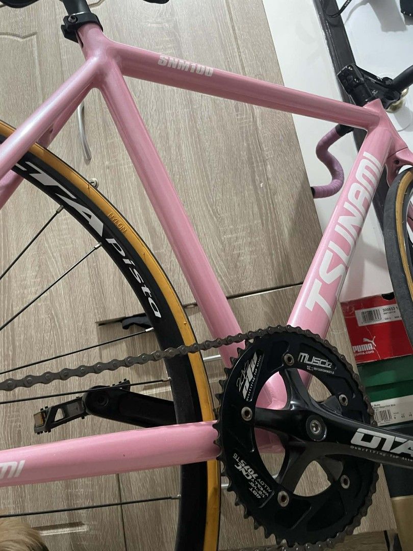 Tsunami SNM100 Fixed Gear Bike (Category: Bicycles) located in Paranaque City. Submitted by newhobbieshop (Listing ID 1430768910, Image 1). Description: Pink Tsunami SNM100 fixed gear bike. Features a black crankset, black handlebars, and a carbon fiber seat. Includes a pair of pedals. The bike has a pink frame and a black wheelset. The tires have a tan sidewall. The handlebars are wrapped in pink tape..