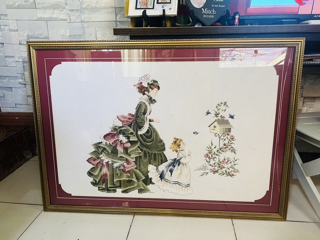 Framed Cross-stitch Embroidery (Category: Frames & Pictures) located in Antipolo. Submitted by murapod2 (Listing ID 1430820956, Image 1). Description: Beautifully framed cross-stitch embroidery featuring a Victorian-era woman and child. The artwork is set against a white background with a maroon border, encased in a gold-colored frame. The embroidery depicts a detailed scene with a birdhouse and floral accents. The piece is ready to hang and woul