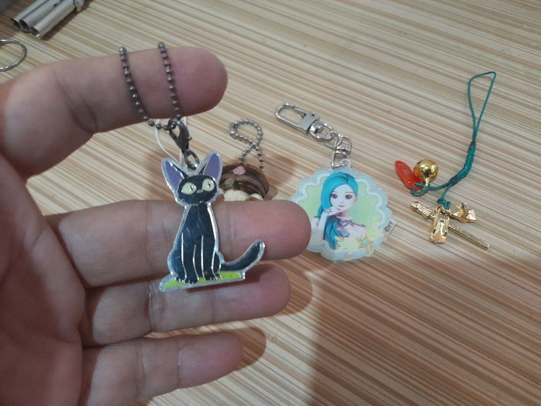 Assorted Keychains (Category: Toys & Games) located in Bocaue. Submitted by marajapansurplus (Listing ID 1431008245, Image 2). Description: Collection of keychains featuring various designs. Includes a black cat, an orange fish, a brown bear-like figure, a girl with blue hair, and a gold-colored cross with a bell. Each keychain has a different attachment style, including a chain, a clasp, and a string. Perfect for adding a touch of personality to