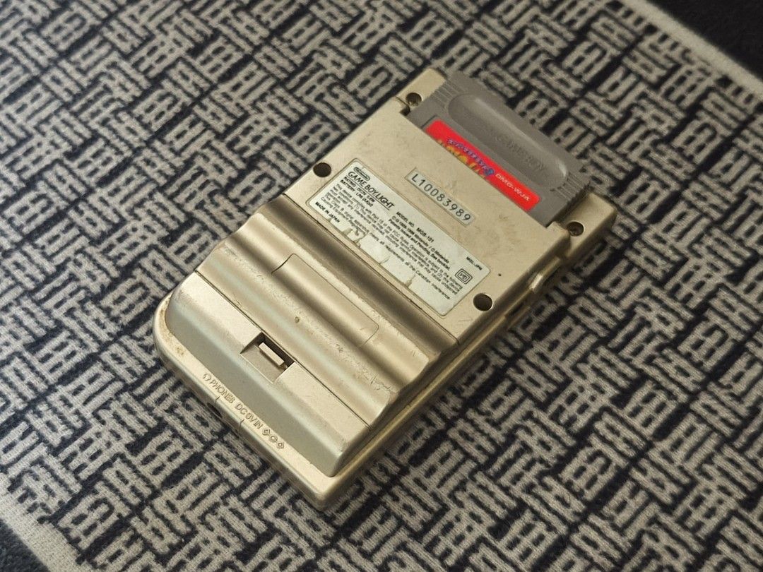Nintendo Game Boy Light (Category: Nintendo) located in Taguig. Submitted by juliestjuly (Listing ID 1437640276, Image 3). Description: Gold-colored Nintendo Game Boy Light. The screen displays a game. The back of the device has a cartridge inserted. The device has a power button, a directional pad, and A and B buttons. The model number is MGB-001. all working with Sounds issue: minor scratches.