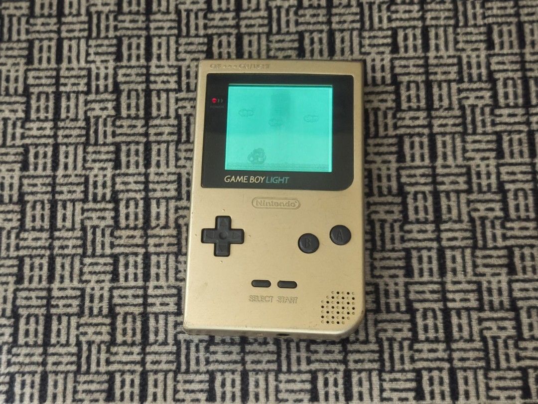 Nintendo Game Boy Light (Category: Nintendo) located in Taguig. Submitted by juliestjuly (Listing ID 1437640276, Image 2). Description: Gold-colored Nintendo Game Boy Light. The screen displays a game. The back of the device has a cartridge inserted. The device has a power button, a directional pad, and A and B buttons. The model number is MGB-001. all working with Sounds issue: minor scratches.