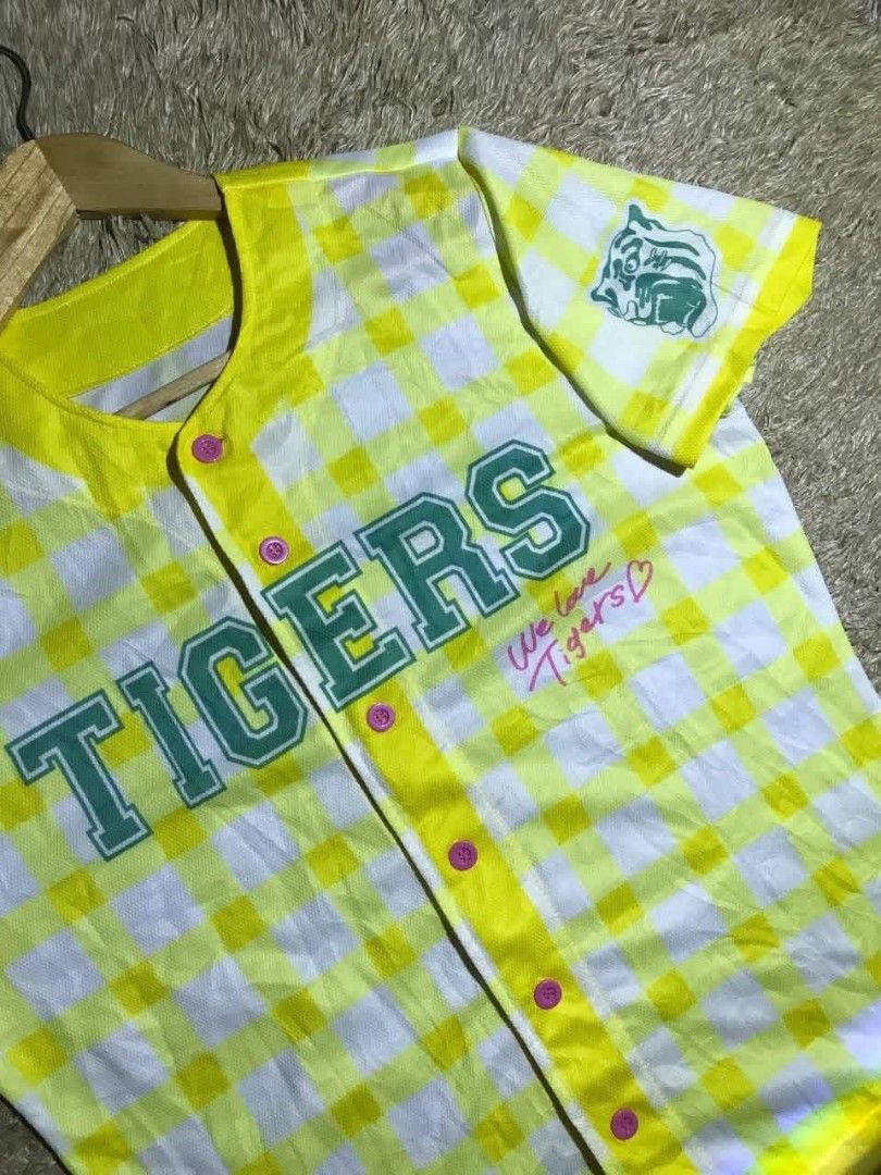 Tigers Baseball Jersey (Category: Tshirts & Polo Shirts ) located in Navotas City. Submitted by jericafits (Listing ID 1438052126, Image 2). Description: - [ ] Tigers Pattern Size: L Dimes: 21 x 26 ₱300 - Yellow and white checkered design - "TIGERS" printed on the front - Button-up style with pink buttons - "We love Tigers" text on the front - Small logo on the sleeve - Short sleeves - Yellow collar.