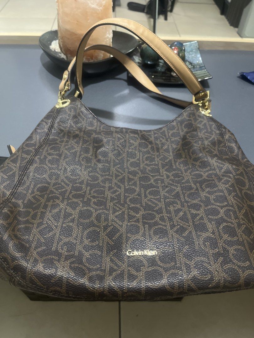 CAlvin Klein (Category: Shoulder Bags) located in Mandaluyong City. Submitted by metrosaka17_ph (Listing ID 1438123876, Image 3). Description: Key FeaturesDesign: Features the signature all-over, repeating “CK” logo monogram pattern.Color Scheme: Dark brown body accented with light tan or camel leather shoulder straps.Hardware: Polished gold-tone metal hardware connecting the handle and zipper compartments.Style: Slouchy hobo structure that rests