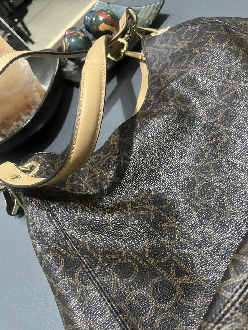 CAlvin Klein (Category: Shoulder Bags) located in Mandaluyong City. Submitted by metrosaka17_ph (Listing ID 1438123876, Image 2). Description: Key FeaturesDesign: Features the signature all-over, repeating “CK” logo monogram pattern.Color Scheme: Dark brown body accented with light tan or camel leather shoulder straps.Hardware: Polished gold-tone metal hardware connecting the handle and zipper compartments.Style: Slouchy hobo structure that rests