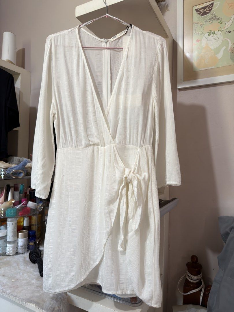 Pull & Bear White Wrap Dress (Category: Dresses) located in Apalit. Submitted by ic_declutter (Listing ID 1435596027, Image 2). Description: White wrap dress from Pull & Bear. Features a V-neckline and long sleeves. The dress has a tie detail at the waist and a flowing skirt. Made in Morocco. Size S/USA S/MEX 26..