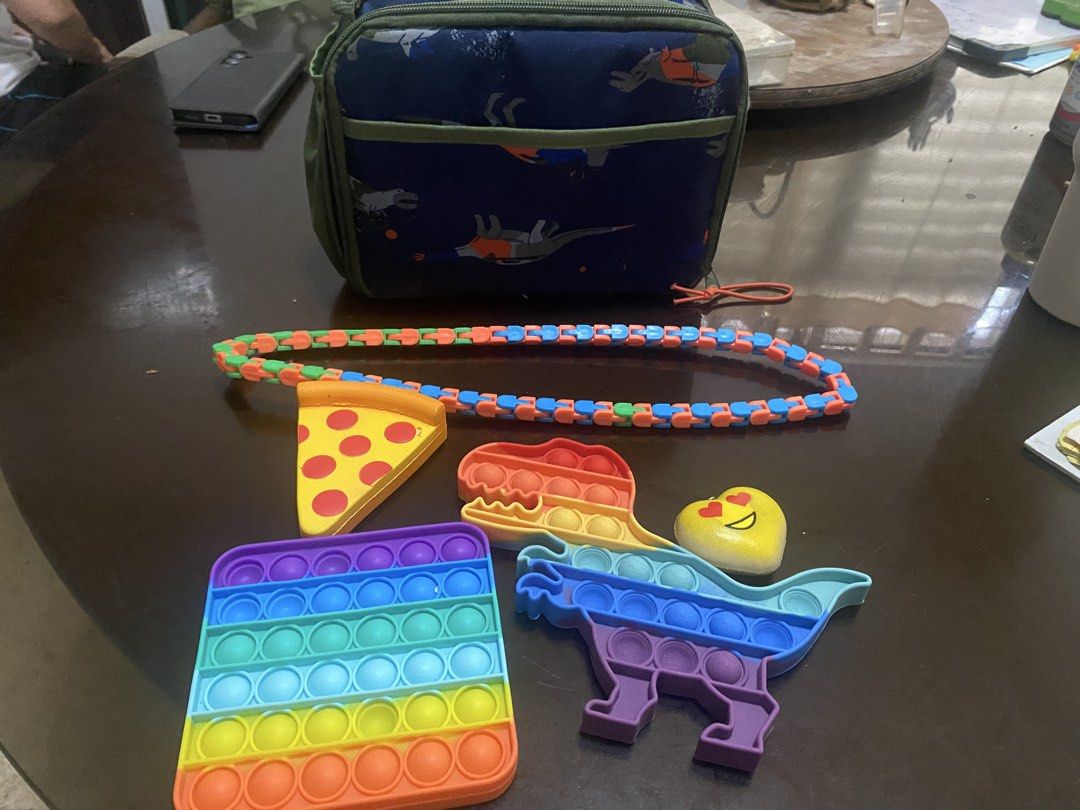BUNDLE Dinosaur Lunch Bag with Fidget Toys (Category: Other Babies Going Out Needs) located in Marikina City. Submitted by sansa16 (Listing ID 1438897787, Image 1). Description: - Dinosaur-themed lunch bag: - Blue exterior with dinosaur prints - Green strap - Side pocket - Zipper closure - Fidget Toys: - Rainbow square pop-it - Pizza slice fidget toy - Heart fidget toy - Dinosaur pop-it - Colorful chain fidget toy.