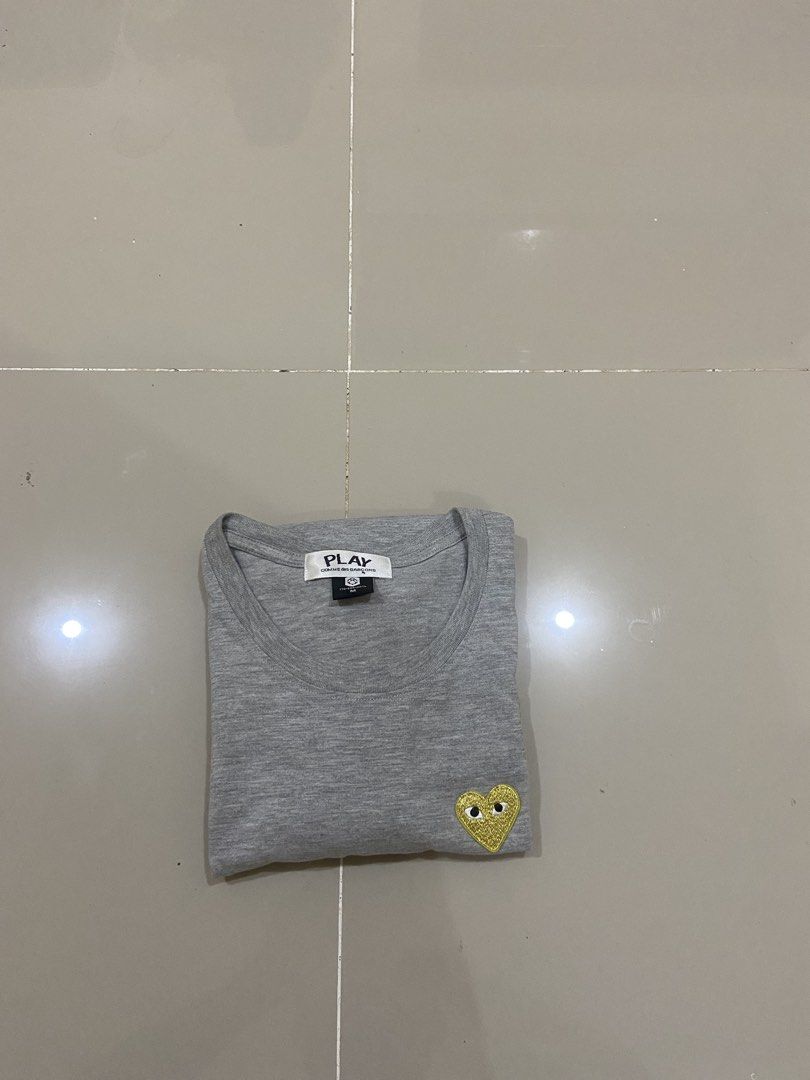 Comme des Garcons Play T-shirt (Category: Shirts) located in Lapu Lapu. Submitted by trip-thrift (Listing ID 1439752920, Image 2). Description: - Gray Comme des Garcons Play t-shirt - Features iconic yellow heart logo - Size: M - Classic crew neck and short sleeves - Authentic brand tag inside collar - Small logo detail on sleeve.