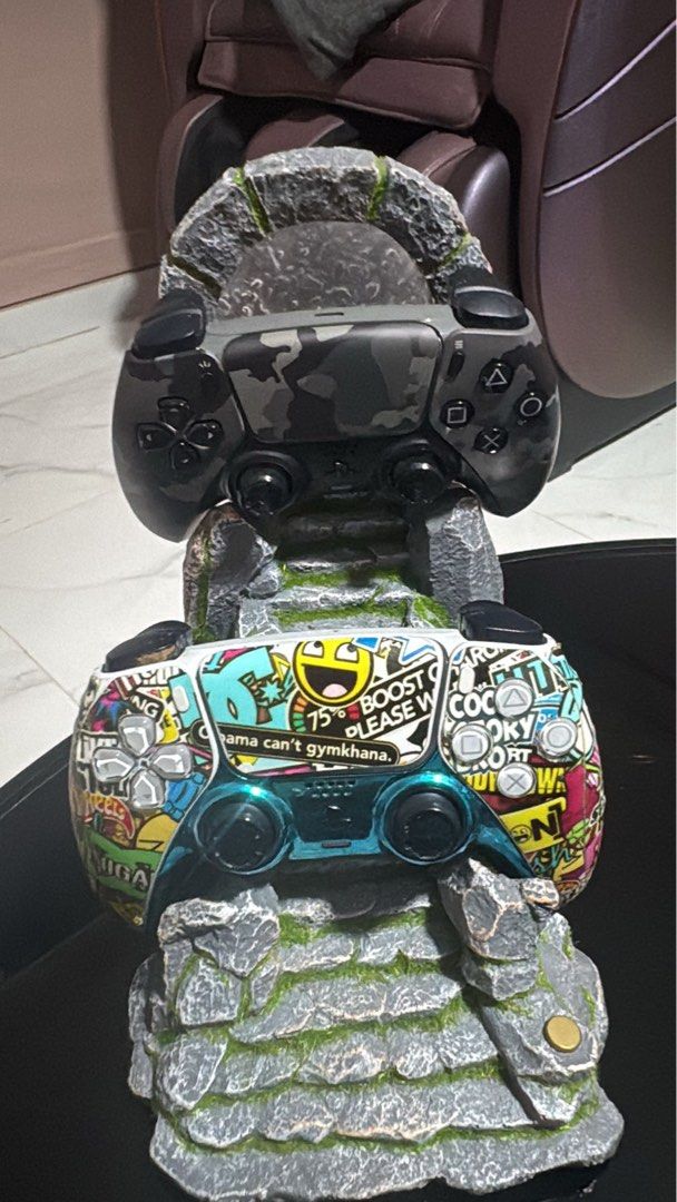 Custom 2 PS5 Controller (Category: Controllers) located in Singapore. Submitted by slayer77777 (Listing ID 1440144358, Image 1). Description: - Unique custom-designed stand for PS5 controllers. - Features a rugged, stone-like appearance with moss accents. - Top controller displays a camouflage skin. - Bottom controller has vibrant decals and meme designs. - Enhances any gamer's setup..