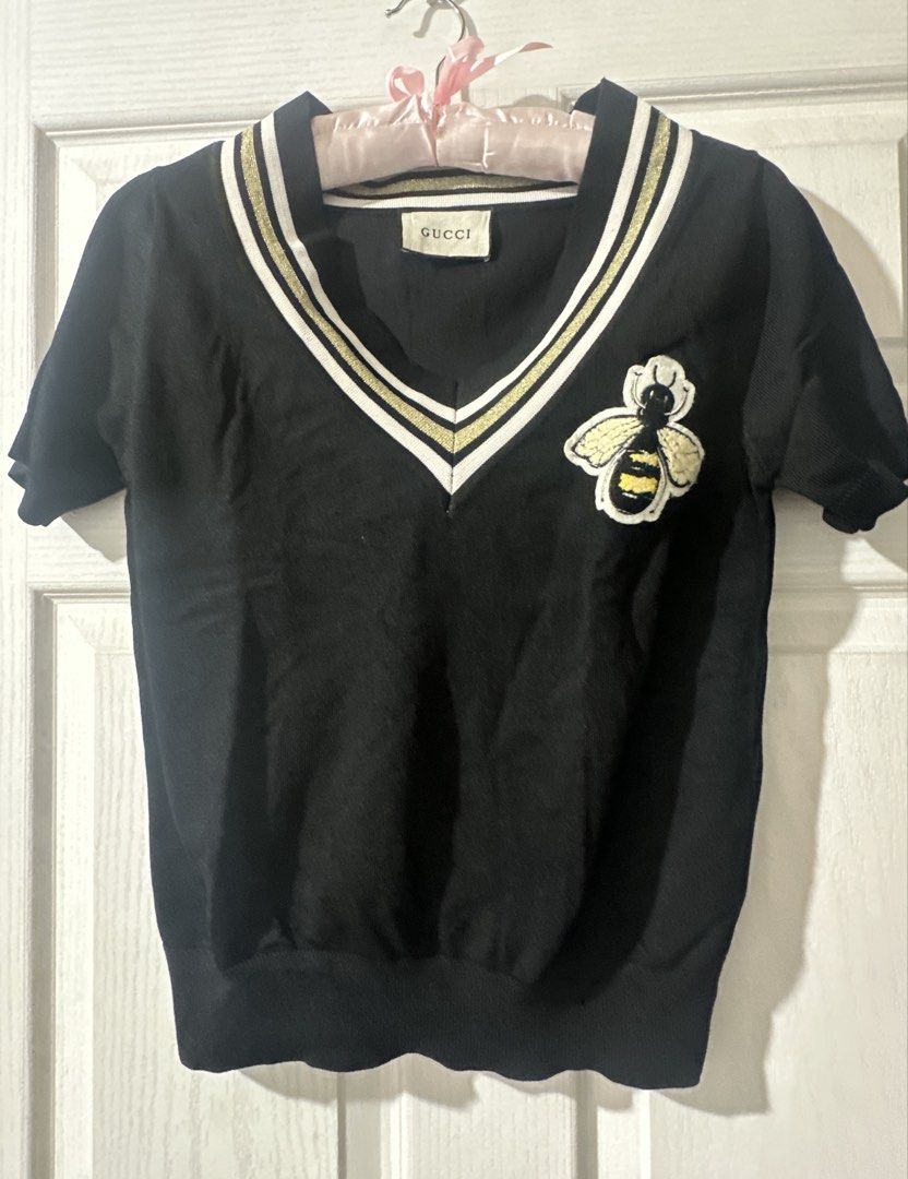 Gucci Black Bee V-Neck Knit Top (Category: Blouses) located in Cabadbaran. Submitted by KsGoodFind (Listing ID 1440329270, Image 1). Description: - Gucci - Color:Black - Ukay source - Style: Short-sleeve knit top - Neckline: V-neck with gold and white striped trim - Detail: Embroidered bee patch on chest - For Casual or dressy wear - Fits Medium.